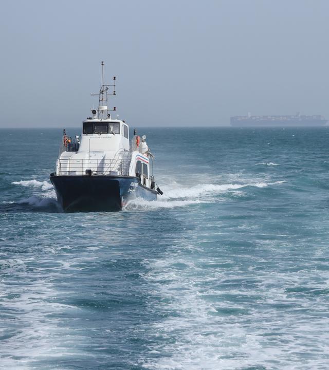 Iran, Bandar Abbas: Ein Patrouillenboot fährt durch das Wasser, während Frachtschiffe in der Straße von Hormuz vor Bandar Abbas, Iran, vor Anker liegen.