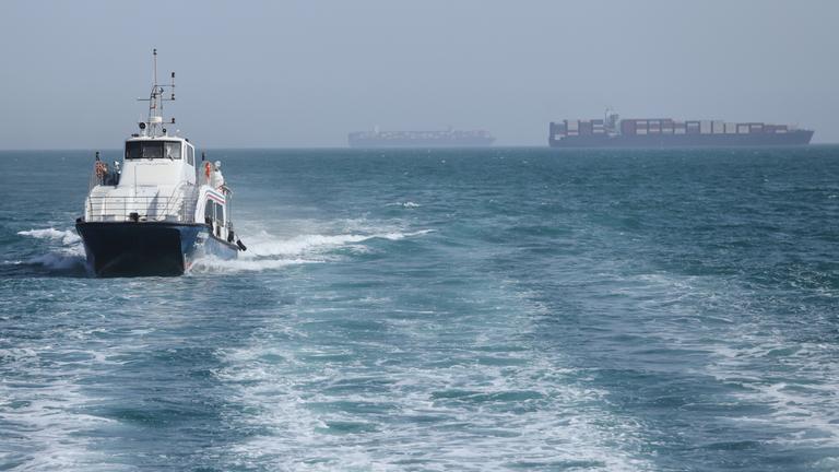 Iran, Bandar Abbas: Ein Patrouillenboot fährt durch das Wasser, während Frachtschiffe in der Straße von Hormuz vor Bandar Abbas, Iran, vor Anker liegen.