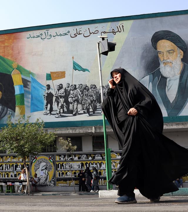 Eine Frau in Burka telefoniert im Laufen. Hinter ihr ist eine Mauer, bemalt unter anderem mit den Gesichtern der Ayatollas Ali Khamenei und Ruhollah Khomeini.