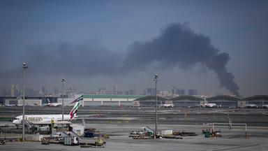 Flugzeuge auf dem Rollfeld des Flughafens Dubai. Im Hintergrund steigt Rauch auf.