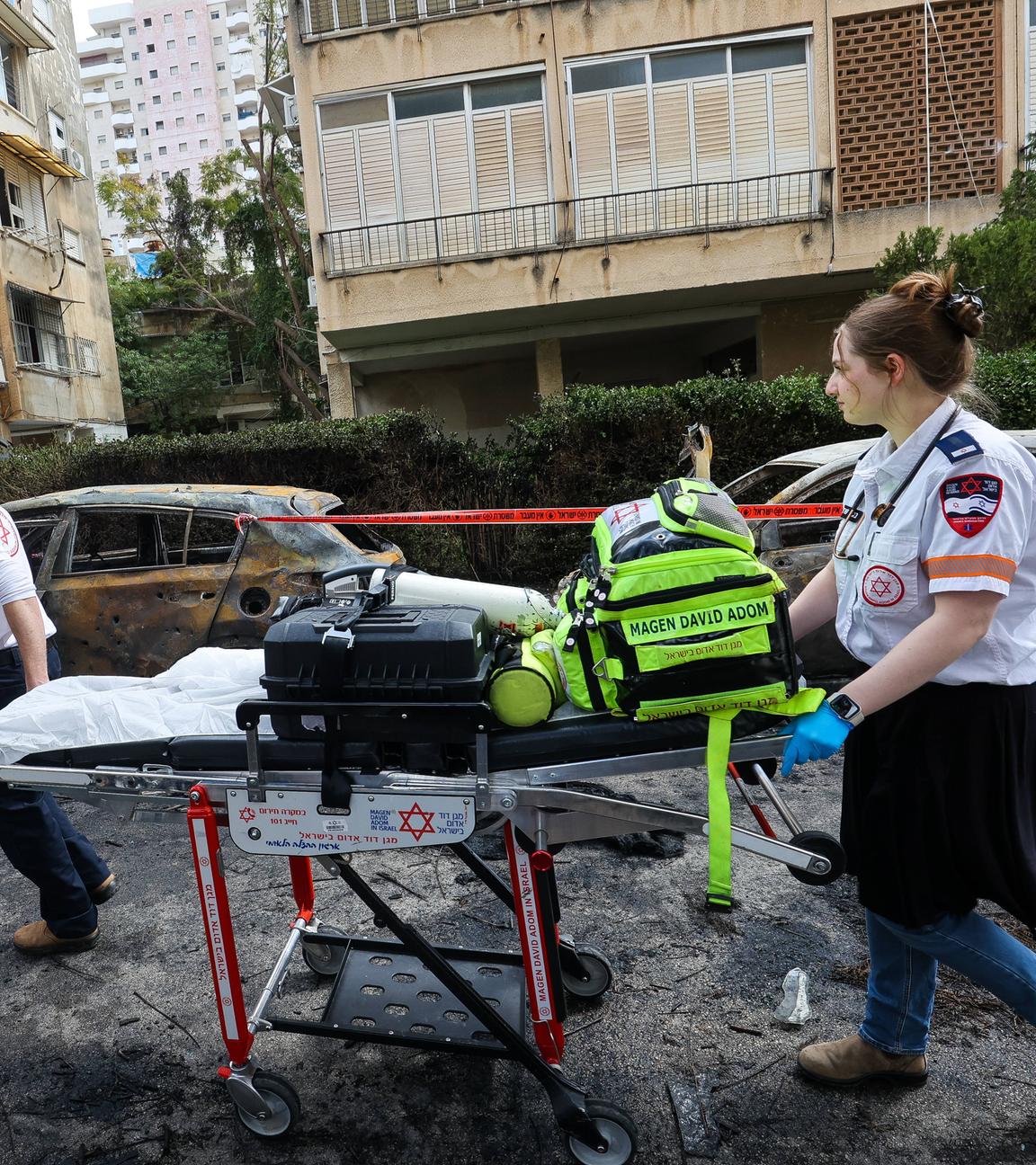 Eine Sanitäterin und ein Sanitäter mit einer Trage im Kriegsgebiet.Im Hintergrund stehen ausgebrannte Autos