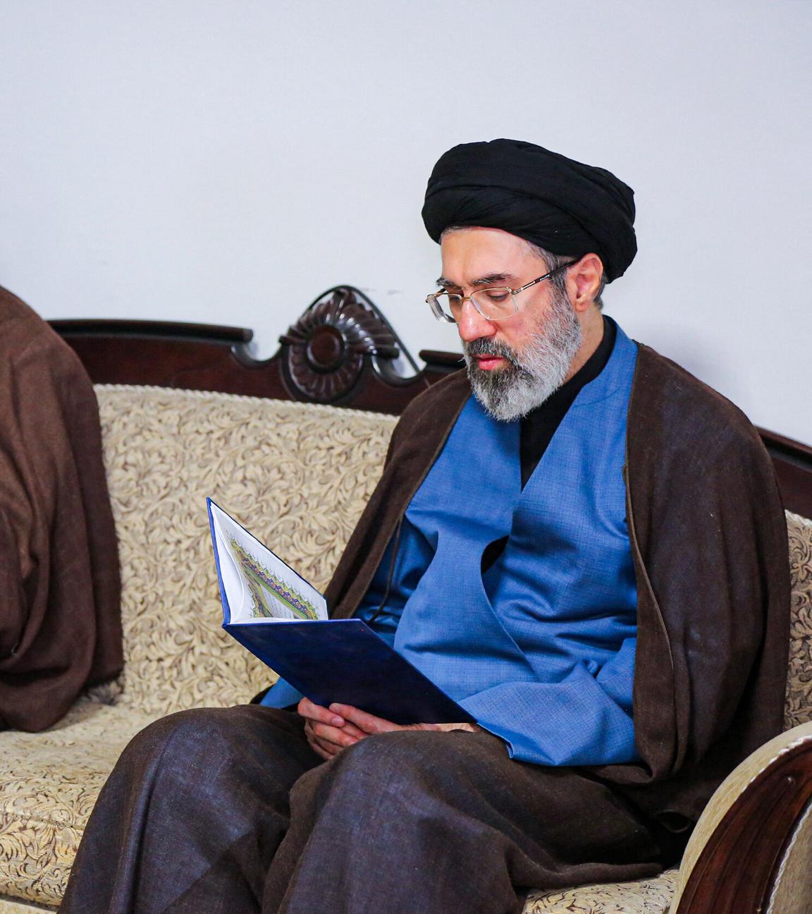 Modschtaba Chamenei sitzt auf einem Sofa und hält ein offenes Buch in der Hand.