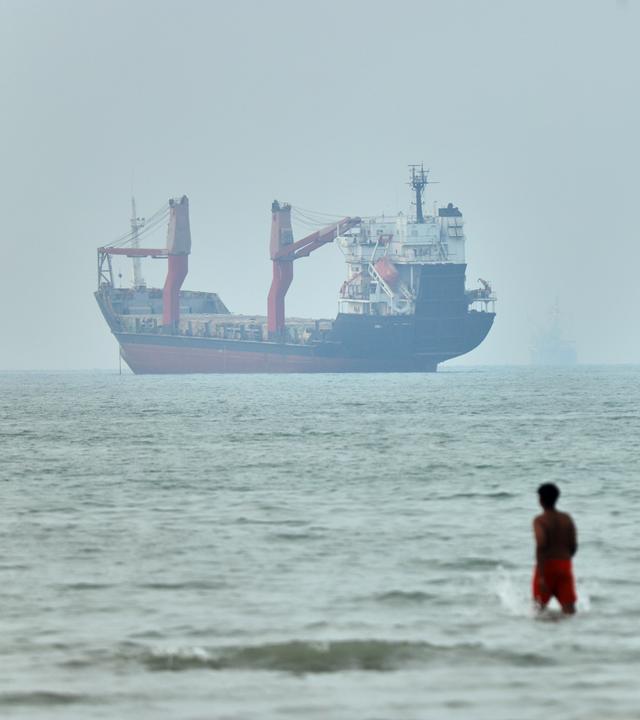 Kinder waten im Wasser. Im Hintergrund liegen vor Anker befindliche Frachtschiffe in der Straße von Hormus.