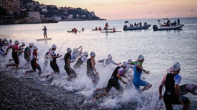 Sportler mit Schwimmkappen in Neopren-Anzügen rennen an einem Kiesstrand ins Meer.