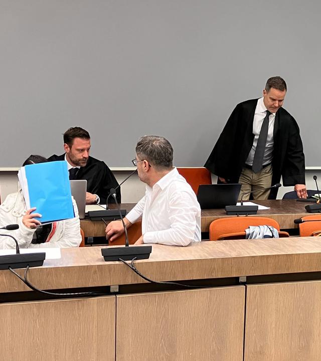 Die wegen Versklavung, Vergewaltigung und Verbrechen gegen die Menschlichkeit angeklagte irakische Frau (2.v.l mit Mappe vor dem Gesicht) sitzt im Saal des Oberlandesgerichts, während ihr mitangeklagter Mann (hinten, 2.v.r) ebenfalls in den Saal kommt.