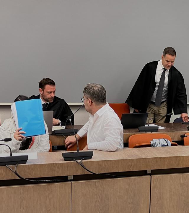 Die wegen Versklavung, Vergewaltigung und Verbrechen gegen die Menschlichkeit angeklagte irakische Frau (2.v.l mit Mappe vor dem Gesicht) sitzt im Saal des Oberlandesgerichts, während ihr mitangeklagter Mann (hinten, 2.v.r) ebenfalls in den Saal kommt.