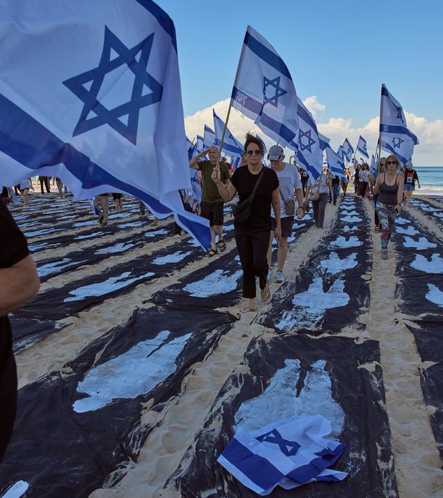 Menschen mit israelischen Flaggen laufen an einem Strand zwischen schwarzen Streifen im Sand, auf die weiß die Umrisse von Personen gemalt sind.