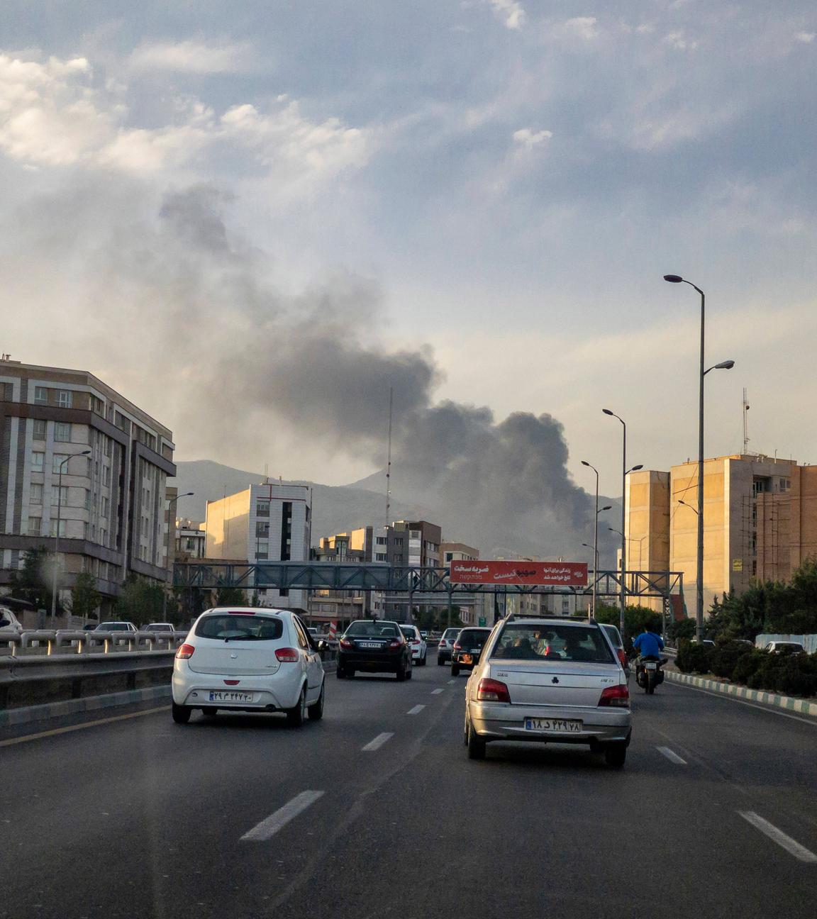 Autos fahren auf einer Straße in Teheran, während im Hintergrund Rauch von einem Angriff aufsteigt.