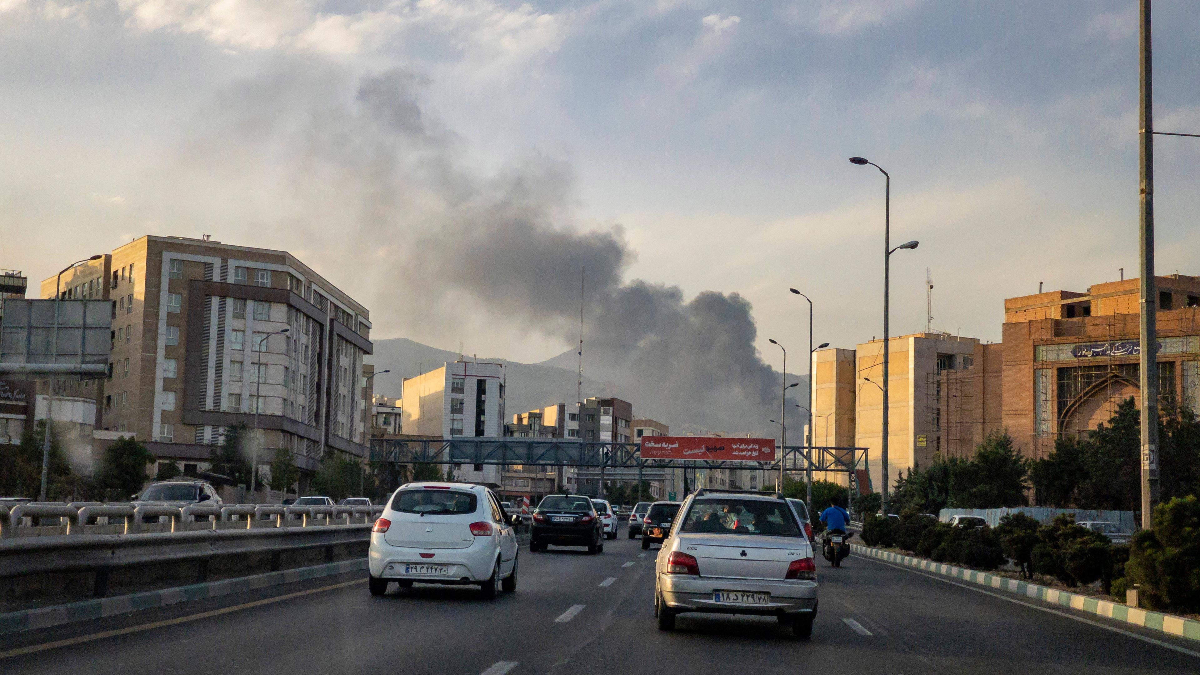 Autos fahren auf einer Straße in Teheran, während im Hintergrund Rauch von einem Angriff aufsteigt.