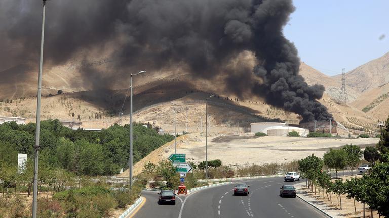 Autos fahren auf einer Autobahn in der Näühe von Teheran, im Hintergrund steigt dunkler Rauch aus einer Ölraffinerie auf, die von israelischen Raketen getroffen wurde.