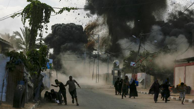 Palästinensische Menschen fliehen vor israelischen Luftangriffen. Im Hintergund steigen dunkle Rauchwolken auf.