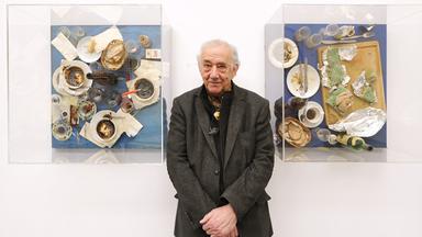 Der Künstler Daniel Spoerri steht zwischen zwei seiner Kunstwerken: der EatArt. Er trägt ein dunklesJackett und ein schwarzes Hemd. Er hält die Hände zusammen vor seinem Bauch. Auf seinen Bildern sind Kaffeetassen, Brot, Aufstriche und Servietten auf einem Tisch zu sehen.