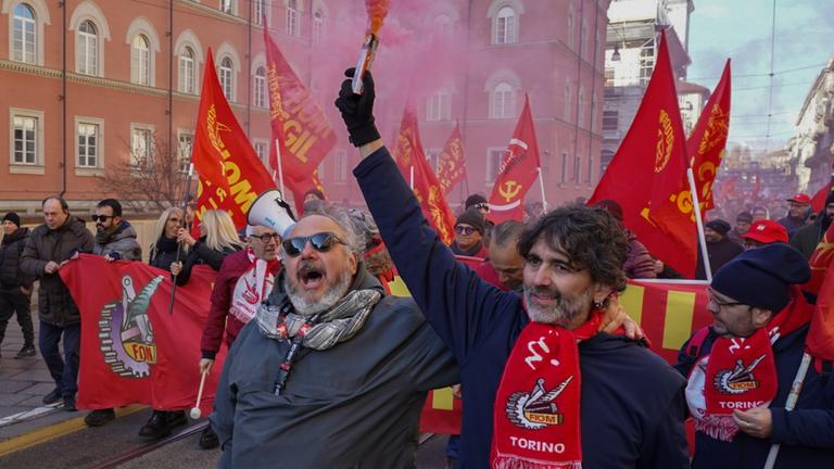 Demonstranten mit Rauchfahnen und Bannern sind in Turin auf einer Straße unterwegs. Zwei Männer sind im Vordergrund zu sehen.