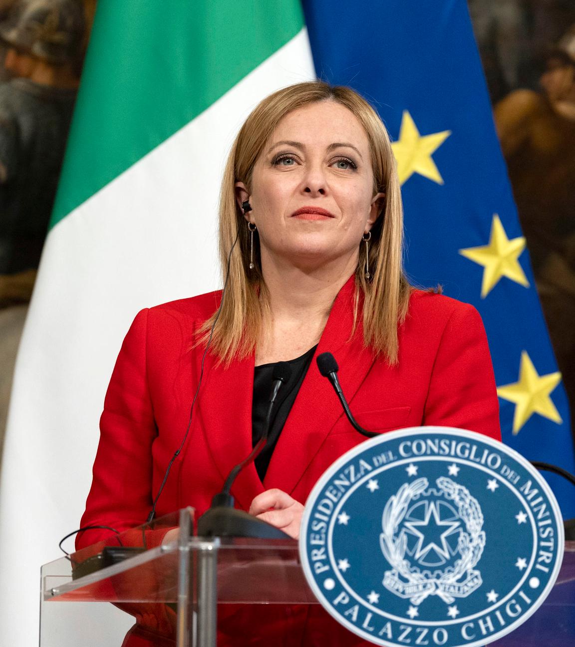 Giorgia Meloni an einem Rednerpult  in einem roten Blazer vor einer Italien und Europaflagge