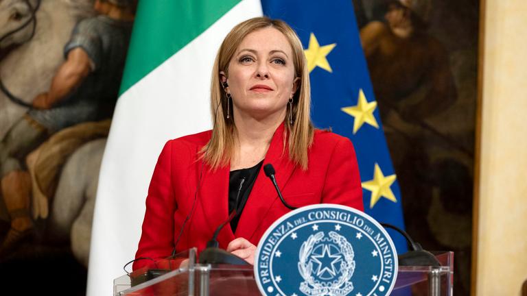 Giorgia Meloni an einem Rednerpult  in einem roten Blazer vor einer Italien und Europaflagge