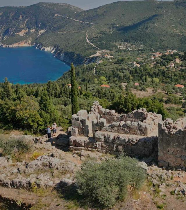 Ruinen stehen auf der Insel Ithaka auf einem Hügel: Dahinter liegt eine Bergkette mit mediterraner Vegetation und eine Meeresbucht.