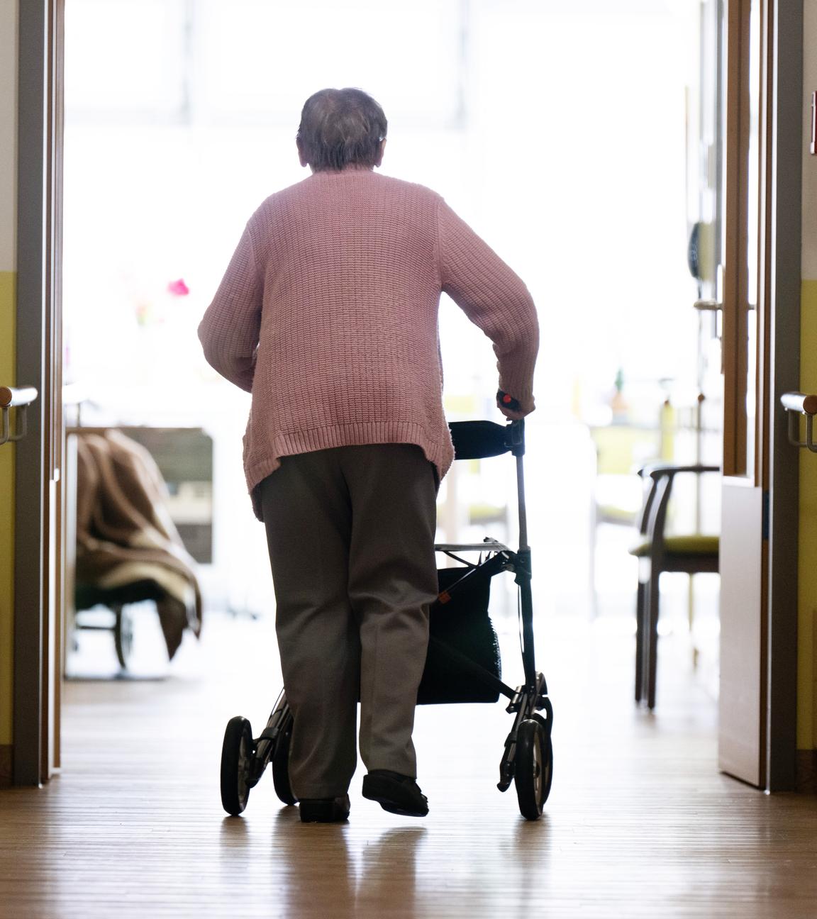 Frau (von hinten zu sehen) läuft mit einem Rollator in einem Altenheim über einen Gang 