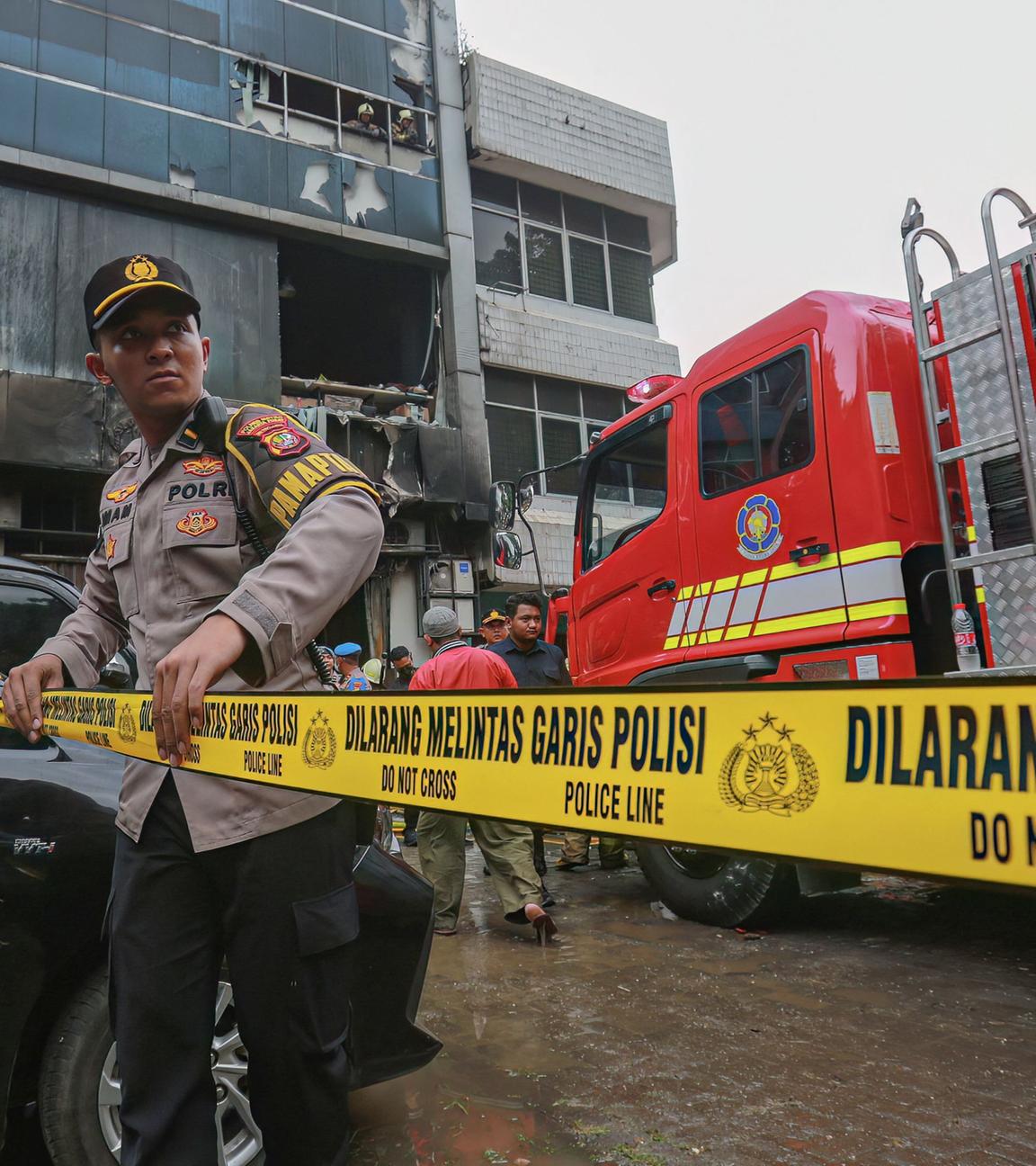 Es ist ein indonesicher Polizist vor einem abgebrannten Haus. Hinter ihm ist ein Feuerwehrfahrzeug. Er sperrt gerade einen bestimmten Bereich mit einem gelben Band ab.