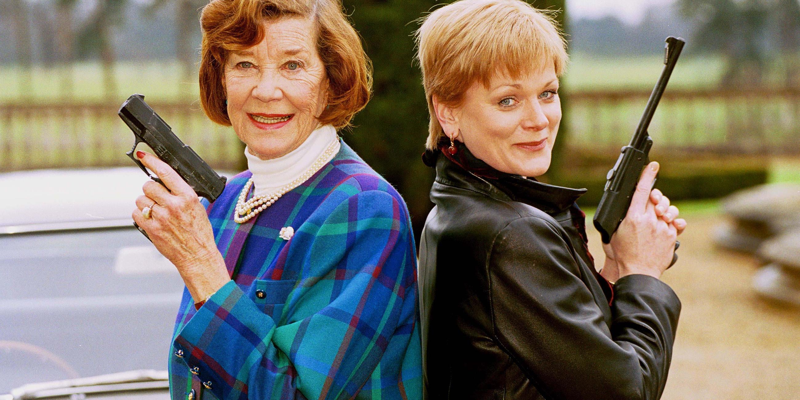 Die Schauspielerinnen Lois Maxwell (l) und Samantha Bond, die beide die Rolle der Miss Moneypenny in verschiedenen James-Bond-Filmen spielten, posieren mit Waffen aus früheren James-Bond-Filmen