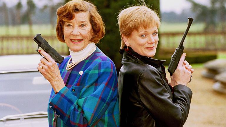 Die Schauspielerinnen Lois Maxwell (l) und Samantha Bond, die beide die Rolle der Miss Moneypenny in verschiedenen James-Bond-Filmen spielten, posieren mit Waffen aus früheren James-Bond-Filmen