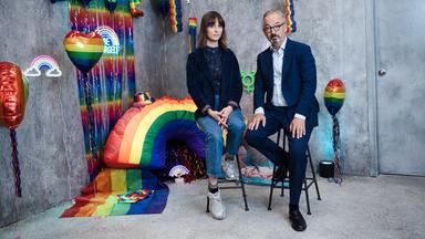 Jan Fleischhauer und Nyke Sławik sitzen auf hohen Hockern vor einer grauen Studiokulisse, die mit Regenbogenfarben und verschiedenen dekorativen Elementen gestaltet ist. Zu sehen sind unter anderem Ballons, kleine Regenbogenflaggen, eine leuchtende Dekoration sowie ein großes Regenbogenobjekt im Hintergrund. Beide blicken in die Kamera. Jan Fleischhauer trägt einen dunklen Anzug mit hellem Hemd, Nyke Sławik ein dunkles Oberteil mit Jeans und Sneakers. Die Szene wirkt wie ein thematisch inszeniertes Studio-Setting.