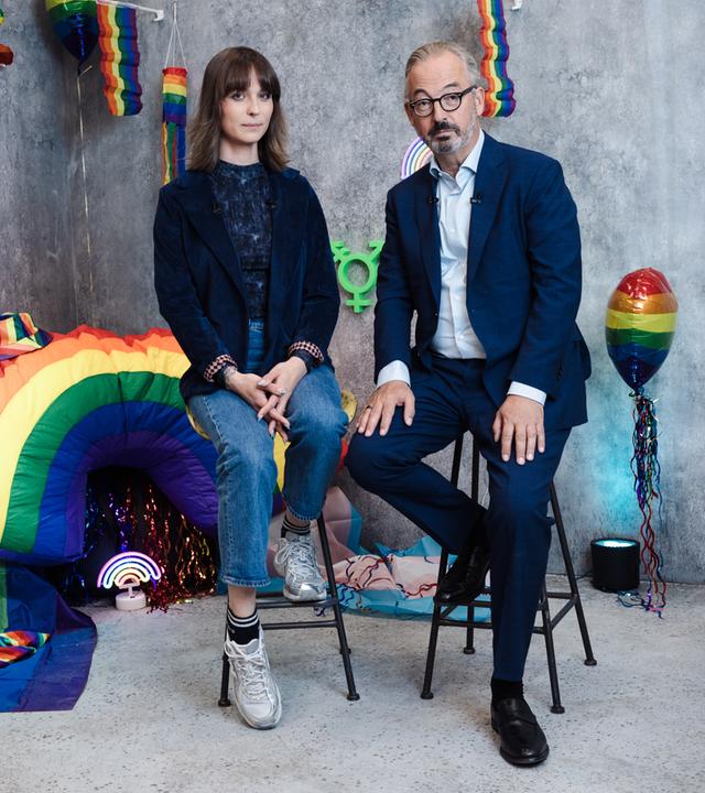 Jan Fleischhauer und Nyke Sławik sitzen auf hohen Hockern vor einer grauen Studiokulisse, die mit Regenbogenfarben und verschiedenen dekorativen Elementen gestaltet ist. Zu sehen sind unter anderem Ballons, kleine Regenbogenflaggen, eine leuchtende Dekoration sowie ein großes Regenbogenobjekt im Hintergrund. Beide blicken in die Kamera. Jan Fleischhauer trägt einen dunklen Anzug mit hellem Hemd, Nyke Sławik ein dunkles Oberteil mit Jeans und Sneakers. Die Szene wirkt wie ein thematisch inszeniertes Studio-Setting.