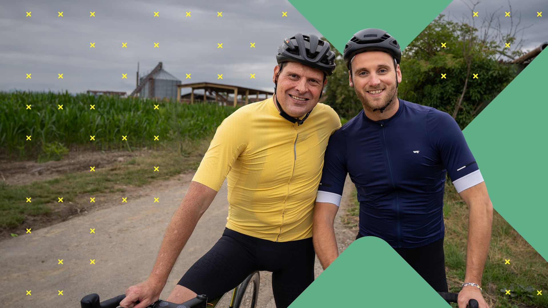 Dr. Leon Windscheid steht lächelnd neben Jan Ullrich auf einem Feldweg. Beide tragen Sportkleidung und Fahrradhelme. Im Hintergrund ist ein Maisfeld und ein grauer Himmel zu sehen.