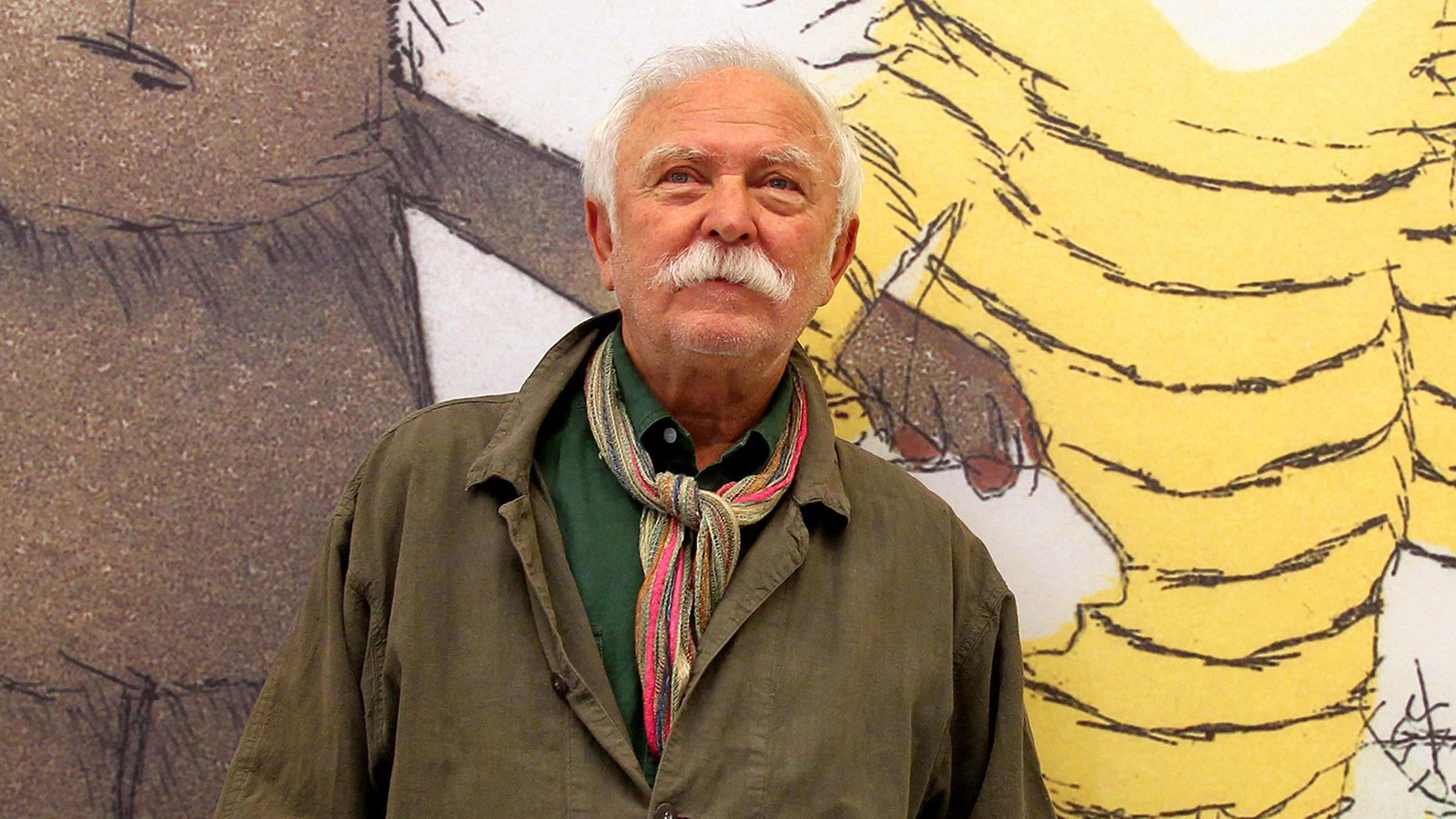 Janosch steht vor einer Wand, auf der seine Figuren abgebildet sind. 