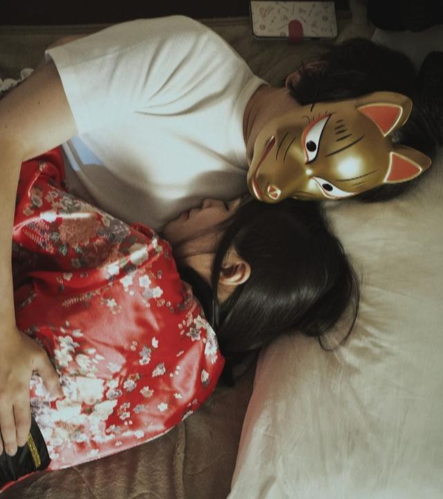 Ein Mann mit goldener Maske und eine Frau im roten Kimono liegen eng umschlungen im Bett.