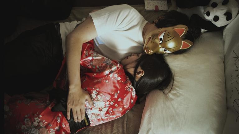 Ein Mann mit goldener Maske und eine Frau im roten Kimono liegen eng umschlungen im Bett.