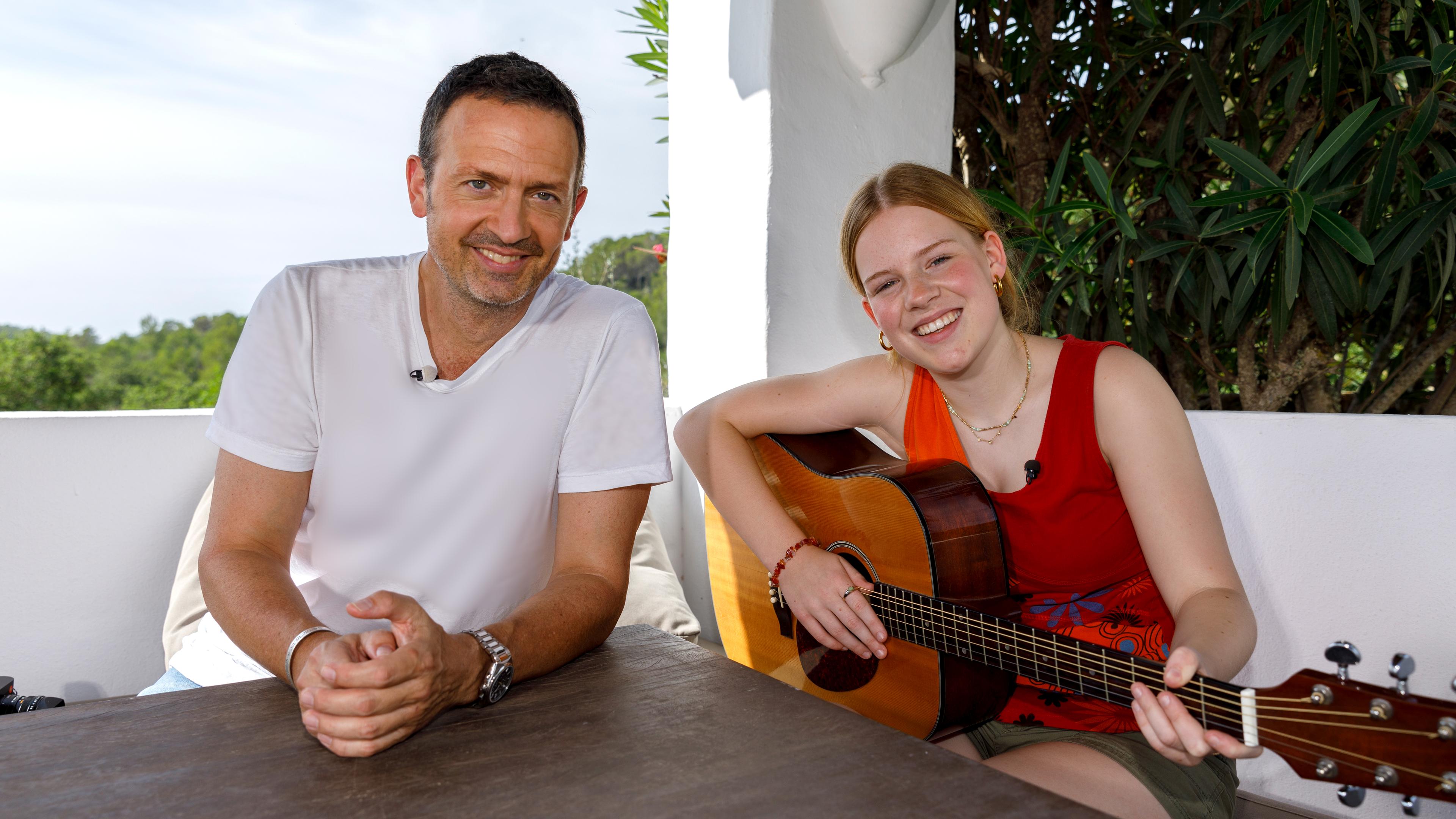 Musikpate Till Brönner und Kandidatin Frida sitzen an einem Tisch im Freien und lächeln in die Kamera. Frida hält dabei eine Gitarre in ihren Händen.