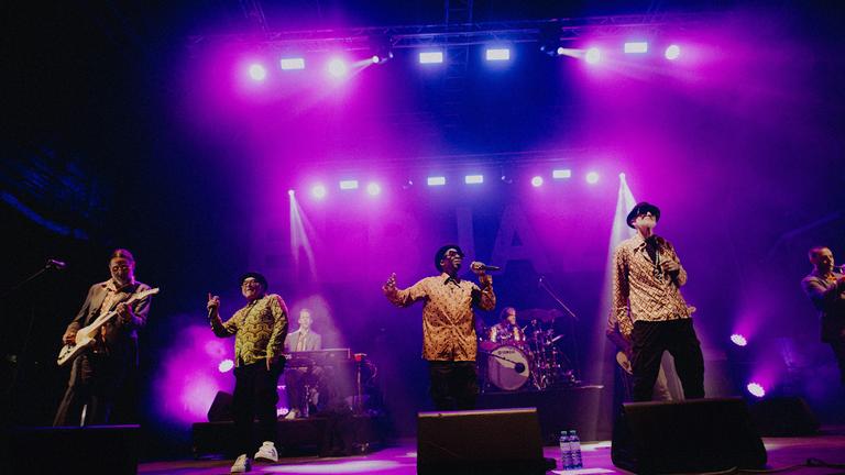 Die Jazzkantinenmitglieder auf der Bühne in Aktion, lila Licht. Vier Sänger in einer Linie in geblümten Hemden und dahinter die Rhythmusgruppe.