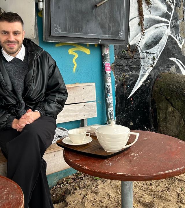Ein junger Mann in dunkler Hose und Lederjacke sitzt auf einer Holzbank vor einem Graffitti-besprächten Gebäude und lächelt in die Kamera.