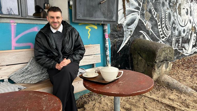 Ein junger Mann in dunkler Hose und Lederjacke sitzt auf einer Holzbank vor einem Graffitti-besprächten Gebäude und lächelt in die Kamera.