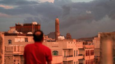 Ein Mann blickt auf ein Minarett, das sich über der Skyline eines Stadtviertels erhebt; aufgenommen bei Sonnenuntergang in Sanaa, der von den Houthis kontrollierten Hauptstadt des Jemen