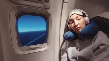 Frau mit Kopfhörern und Nackenkissen sitzt im Flugzeug und schläft.