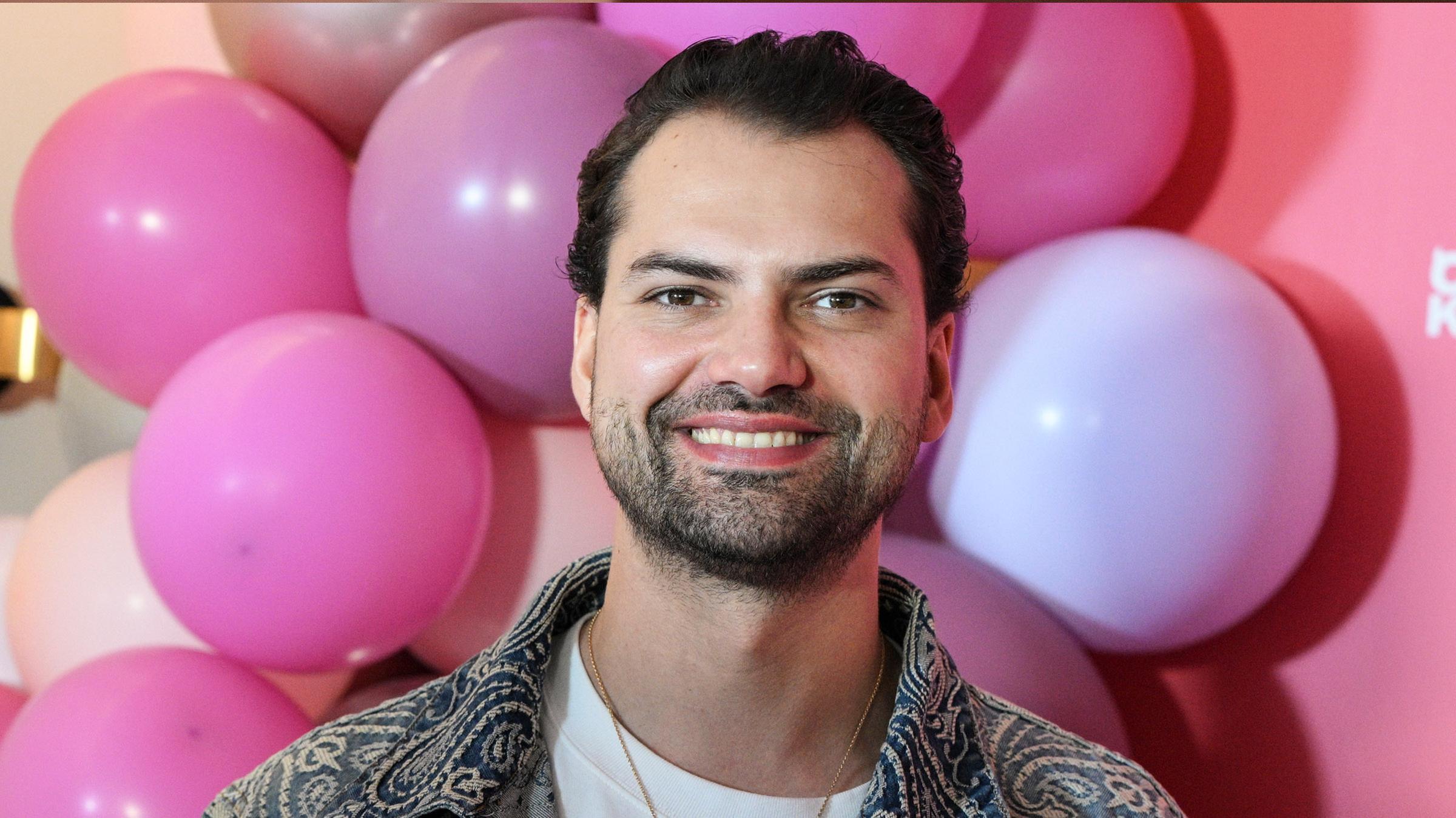 Jimi Blue Ochsenknecht bei einem Pressetermin. Im Hintergrund sind pinke und rosafarbende Luftballons.