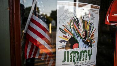 Die US-Amerikanische Flagge hinter einer Schaufensterscheibe. Daneben ein Plakat zu Ehren des hundertsten Geburtstag vom ehemaligen Präsidenten der Vereinigten Staaten: Jimmy Carter.