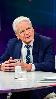 Joachim Gauck sitzt links im Bild an einem hellen, glänzenden Studiotisch und blickt zu Maybrit Illner. Er lehnt sich leicht nach vorn und führt seine Hände über der Tischplatte zusammen. Gauck trägt einen dunkelblauen Anzug, ein weißes Hemd und eine blau-grün gestreifte Krawatte. Illner ist rechts im Vordergrund von hinten zu sehen und trägt einen pinkfarbenen Blazer. Auf dem Tisch stehen Wassergläser und eine Glaskaraffe.