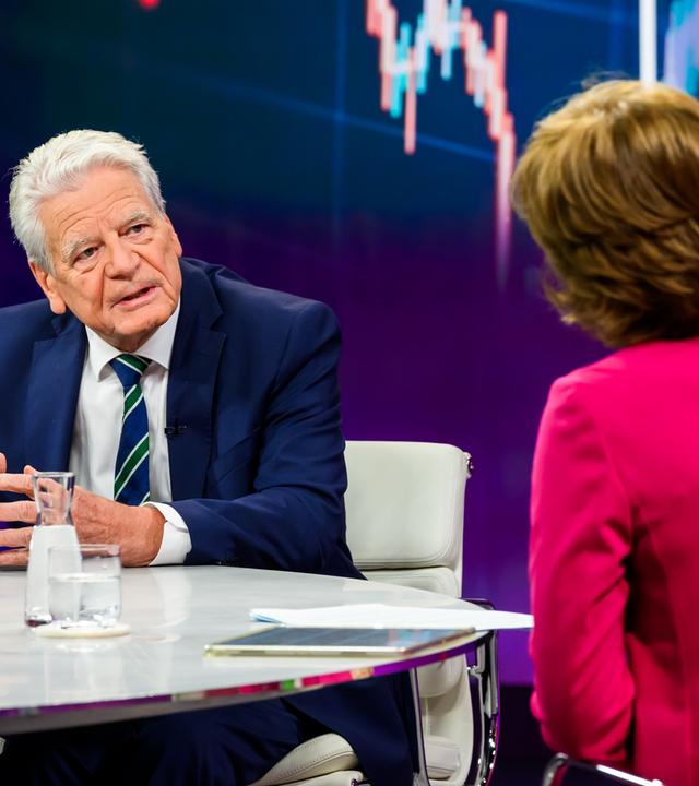 Joachim Gauck sitzt links im Bild an einem hellen, glänzenden Studiotisch und blickt zu Maybrit Illner. Er lehnt sich leicht nach vorn und führt seine Hände über der Tischplatte zusammen. Gauck trägt einen dunkelblauen Anzug, ein weißes Hemd und eine blau-grün gestreifte Krawatte. Illner ist rechts im Vordergrund von hinten zu sehen und trägt einen pinkfarbenen Blazer. Auf dem Tisch stehen Wassergläser und eine Glaskaraffe.