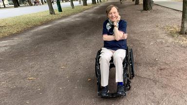 Mann sitzt im Rollstuhl auf einer Allee und lächelt.