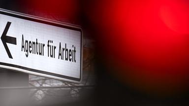 Eine rote Ampel leuchtet vor einem Schild, das auf die Agentur für Arbeit hinweist. (Archiv)