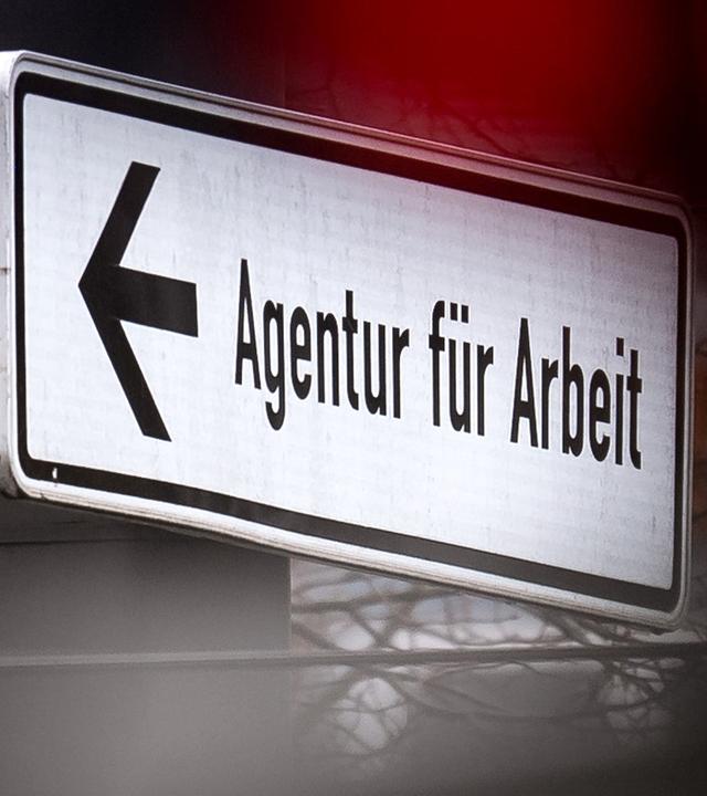 Eine rote Ampel leuchtet vor einem Schild, das auf die Agentur für Arbeit hinweist. (Archiv)