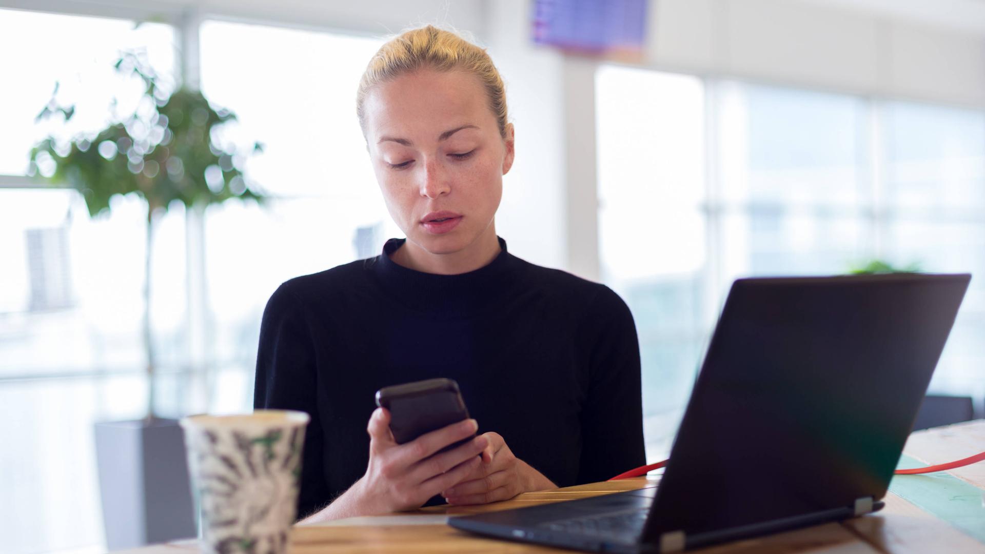 Eine Frau sitzt vor einem Laptop und hat gleichzeitig ein Smaprtphone in der Hand, auf das sie blickt. 