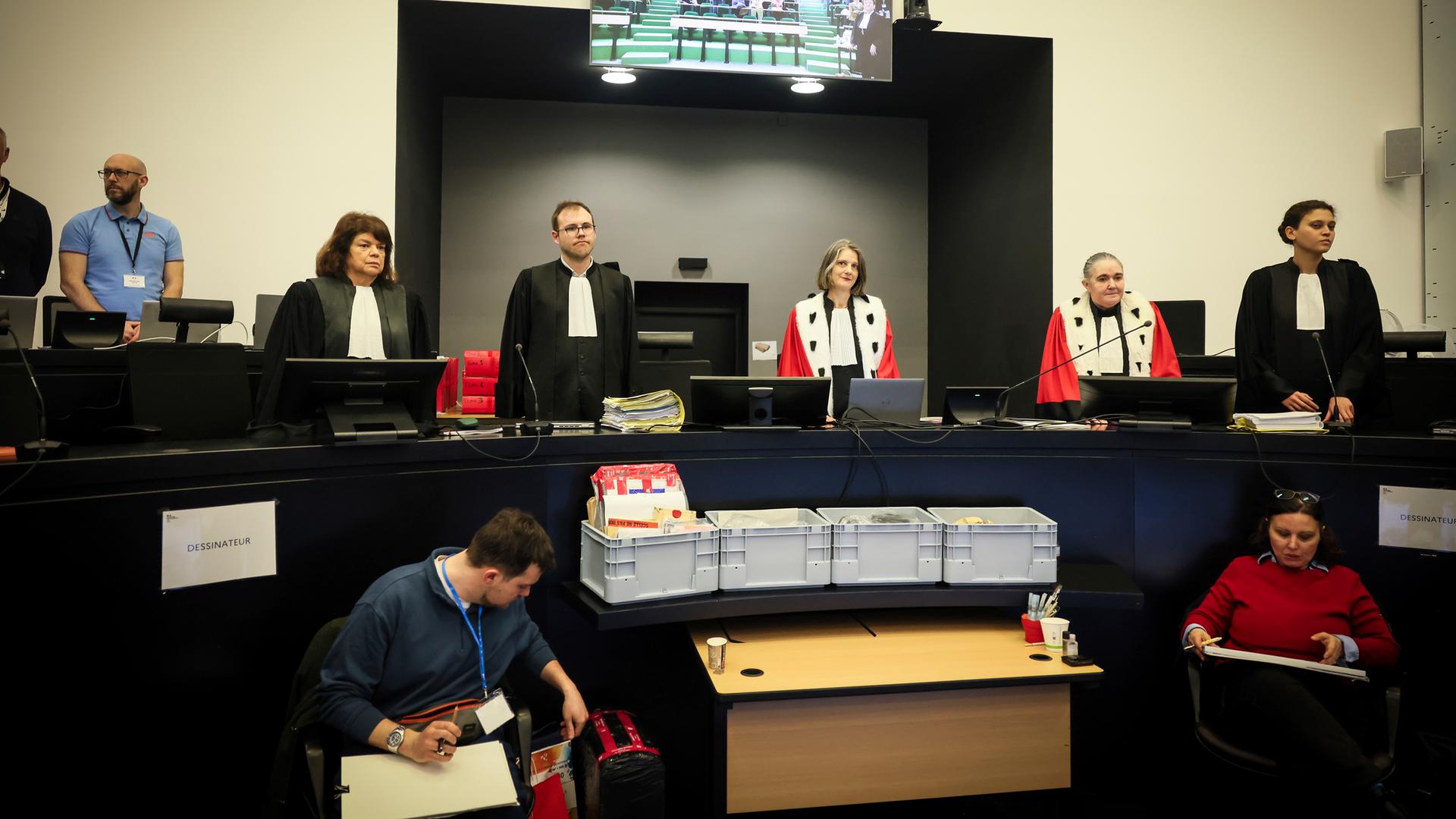 In einem Gerichtssaal in Frankreich stehen der Gerichtspräsident und die Anwälte hinter ihren Tischen. Vor ihnen stehen kistenweise Akten.