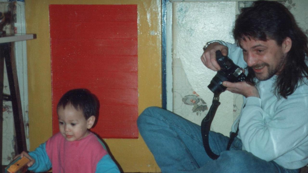 Ein Baby sitzt neben einem Mann, der eine Kamera hält; beide lächeln.