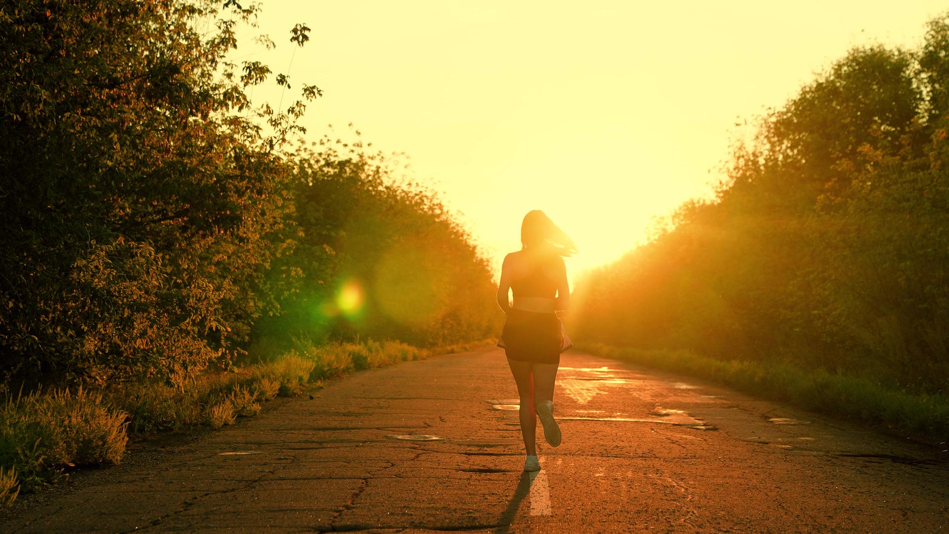 Frau joggt auf einer von Büschen gesäumten Straße in den Sonnenuntergang.