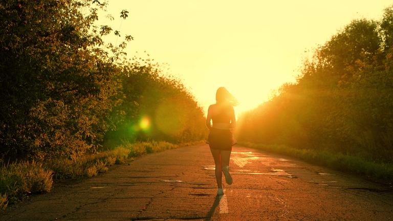 Frau joggt auf einer von Büschen gesäumten Straße in den Sonnenuntergang.