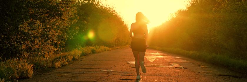 Frau joggt auf einer von Büschen gesäumten Straße in den Sonnenuntergang.