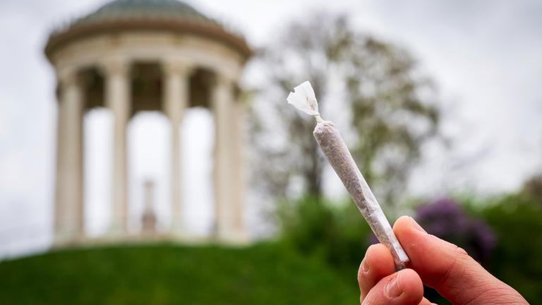 Ein Mann hält einen Joint vor der Kulisse des Monopteros im Englischen Garten